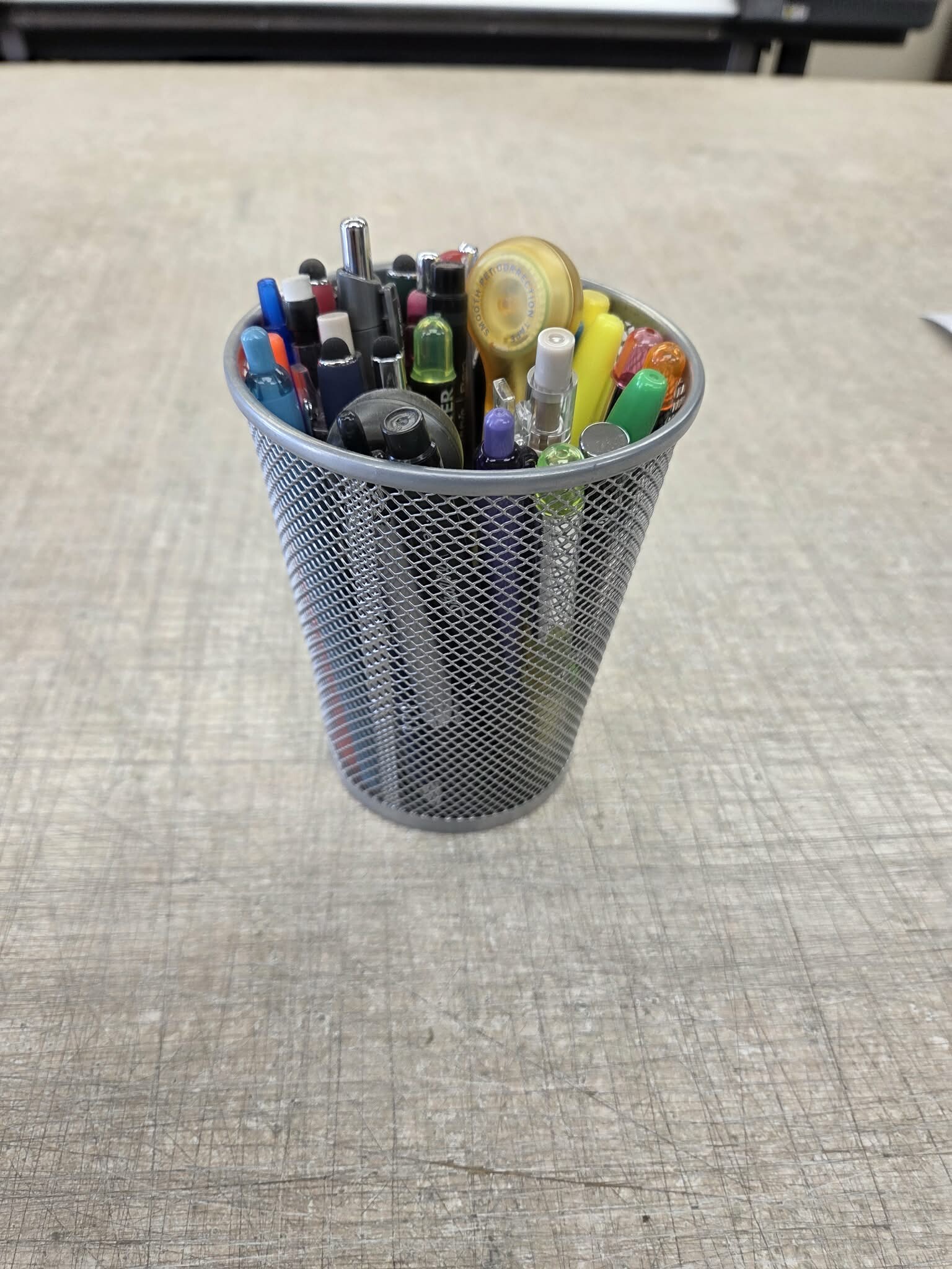 A plain black mesh pencil cup filled with pens and pencils